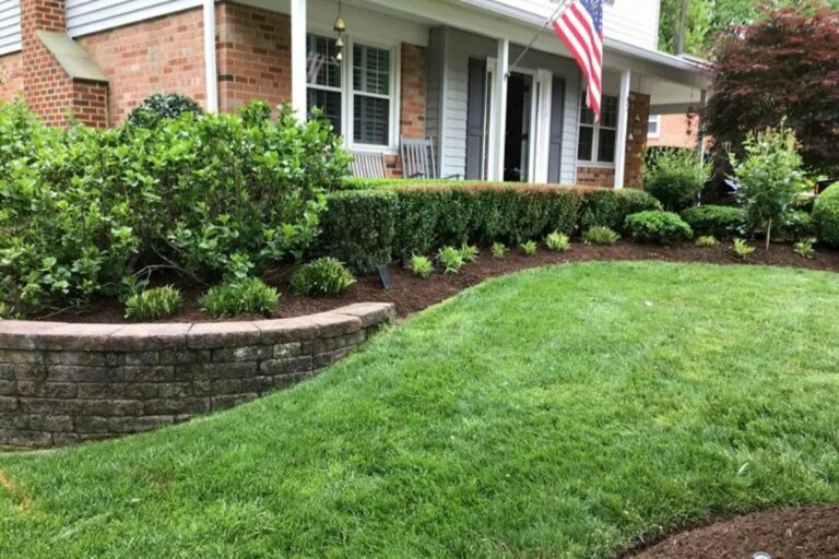 A front yard landscape bed with shrubs, mulch, lawn, and a brick home