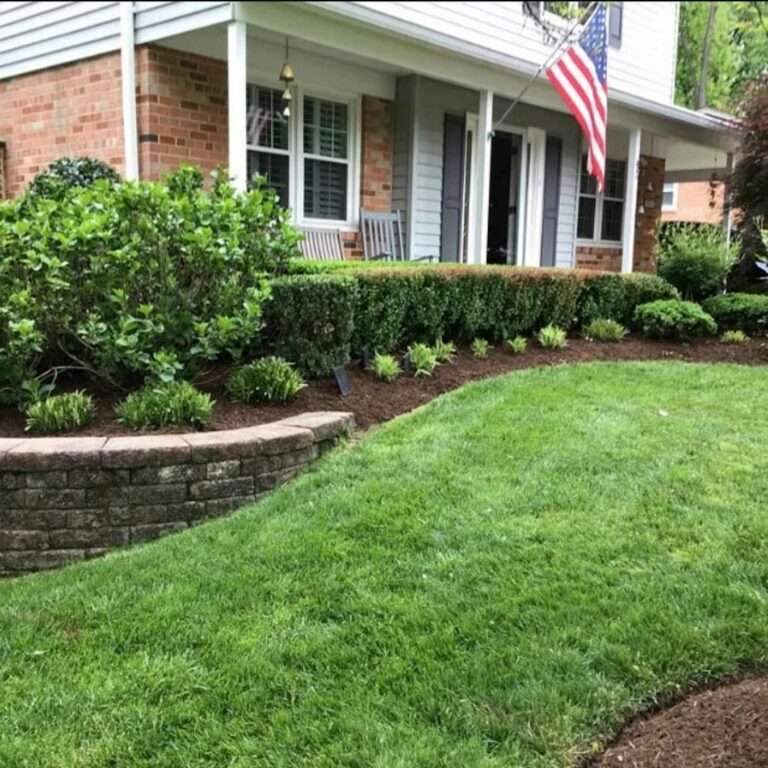 A front yard landscape bed with shrubs, mulch, lawn, and a brick home